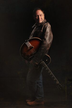 Image of DC Scott wearing leather jacket with guitar slung down to floor